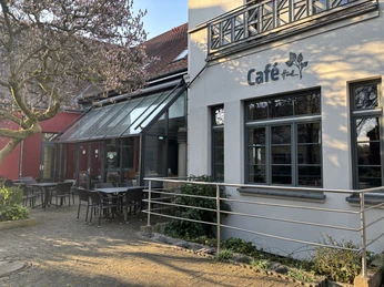 Herford-Café Hudl-Pro Herford GmbH (90).jpeg Außenansicht des Café Hudl mit Glasfassade und Sitzgelegenheiten im Freien bei sonnigem Wetter.