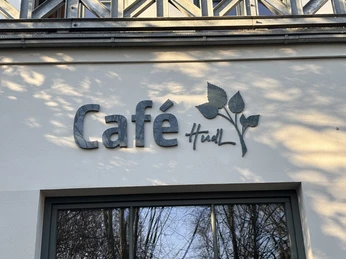 Herford-Café Hudl-Pro Herford GmbH (91).jpeg Metallschriftzug "Café Hudl" an weißer Fassade, umgeben von Schattenspielen der Bäume.