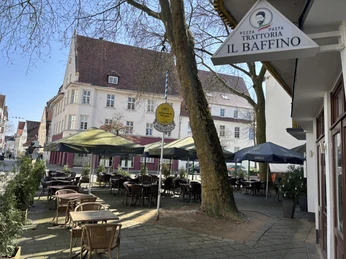 Herford-Trattoria il Baffino-Pro Herford GmbH (77).jpeg Außenterrasse der Trattoria Il Baffino in Herford mit Holztischen und Sonnenschirmen unter Bäumen.