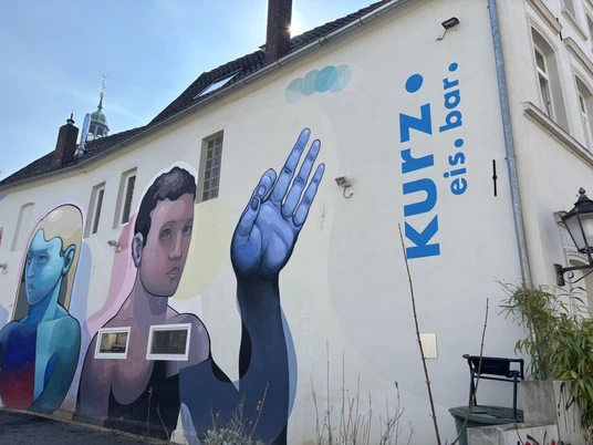 Herford-Kurz Eis Bar-Pro Herford GmbH (59).jpeg Großflächiges Wandgemälde an der Außenwand einer weißen Eisbar mit blauen und rosa Farbtönen.