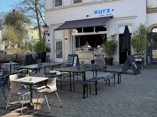 Herford-Kurz Eis Bar-Pro Herford GmbH (60).jpeg Eisbar mit Außensitzbereich, moderner Einrichtung und einladendem Ambiente unter blauem Himmel.