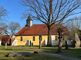 Gelbe Kirche mit rotem Dach und Turm, umgeben von Bäumen auf grünem Rasen unter blauem Himmel.