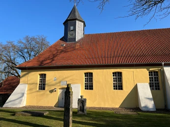 Gelb gestrichene Backsteinkirche mit rotem Schindeldach, Kirchenuhr und angrenzendem Friedhof.