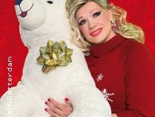 Eine Person in rotem Pullover posiert lächelnd neben einem großen weißen Plüsch-Eisbären. Der Hintergrund ist rot.