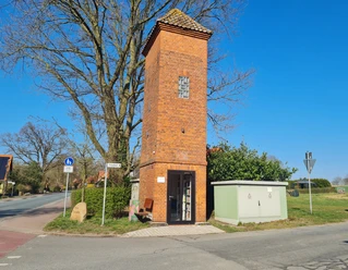 Trafoturm Brokeloh Roter Backsteinturm mit bogenförmigen Fenstern, umgeben von einem Stromkasten und einer Sitzbank an der Straße.