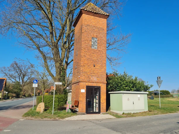 Trafoturm Brokeloh Roter Backsteinturm mit bogenförmigen Fenstern, umgeben von einem Stromkasten und einer Sitzbank an der Straße.