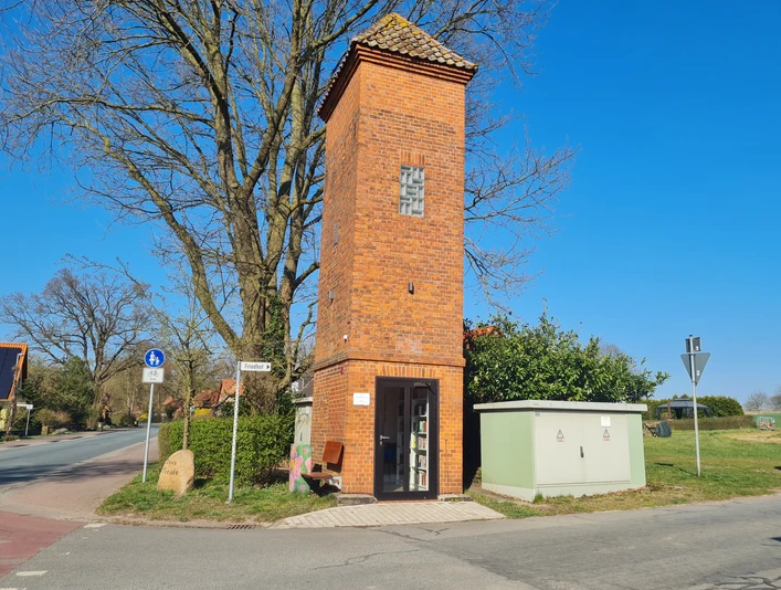 Trafoturm Brokeloh Roter Backsteinturm mit bogenförmigen Fenstern, umgeben von einem Stromkasten und einer Sitzbank an der Straße.