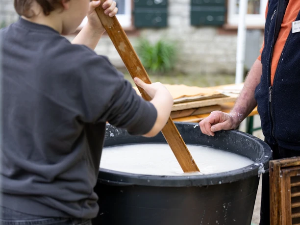 Papier- und Fairmarkt Zwei Personen rühren eine milchige Flüssigkeit in einem großen Kunststoffbehälter um, vermutlich beim Papiermachen.