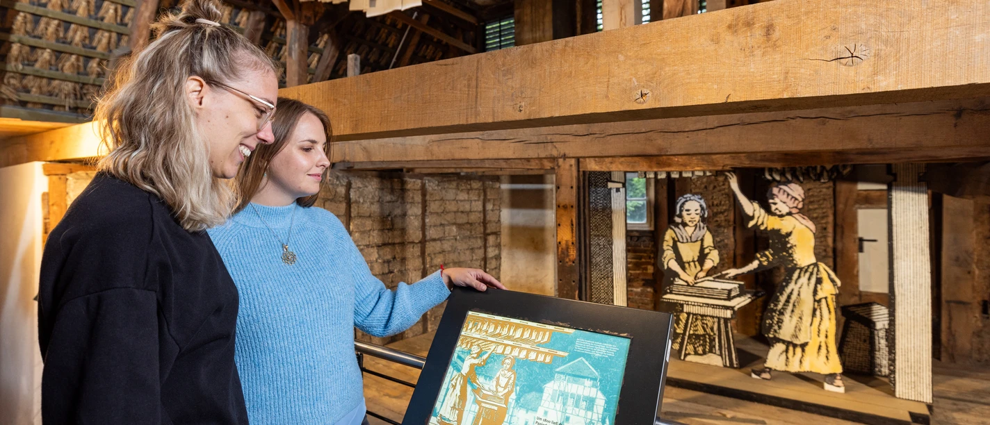 LVR-Industriemuseum Papiermühle Alte Dombach Zwei Frauen betrachten eine historische Ausstellung in einem hölzernen, rustikalen Gebäude.