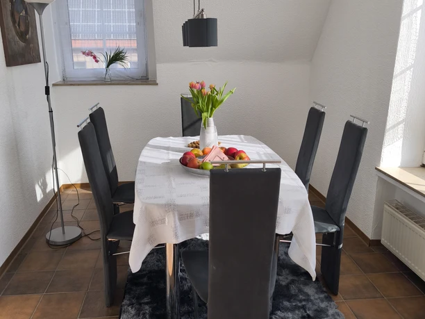 Essbereich.jpg Moderner Essbereich mit Tisch, sechs Stühlen, Tulpenvase und Obstschale vor einem Fenster.