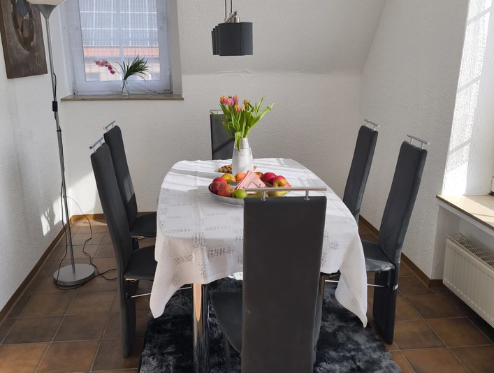 Essbereich.jpg Moderner Essbereich mit Tisch, sechs Stühlen, Tulpenvase und Obstschale vor einem Fenster.