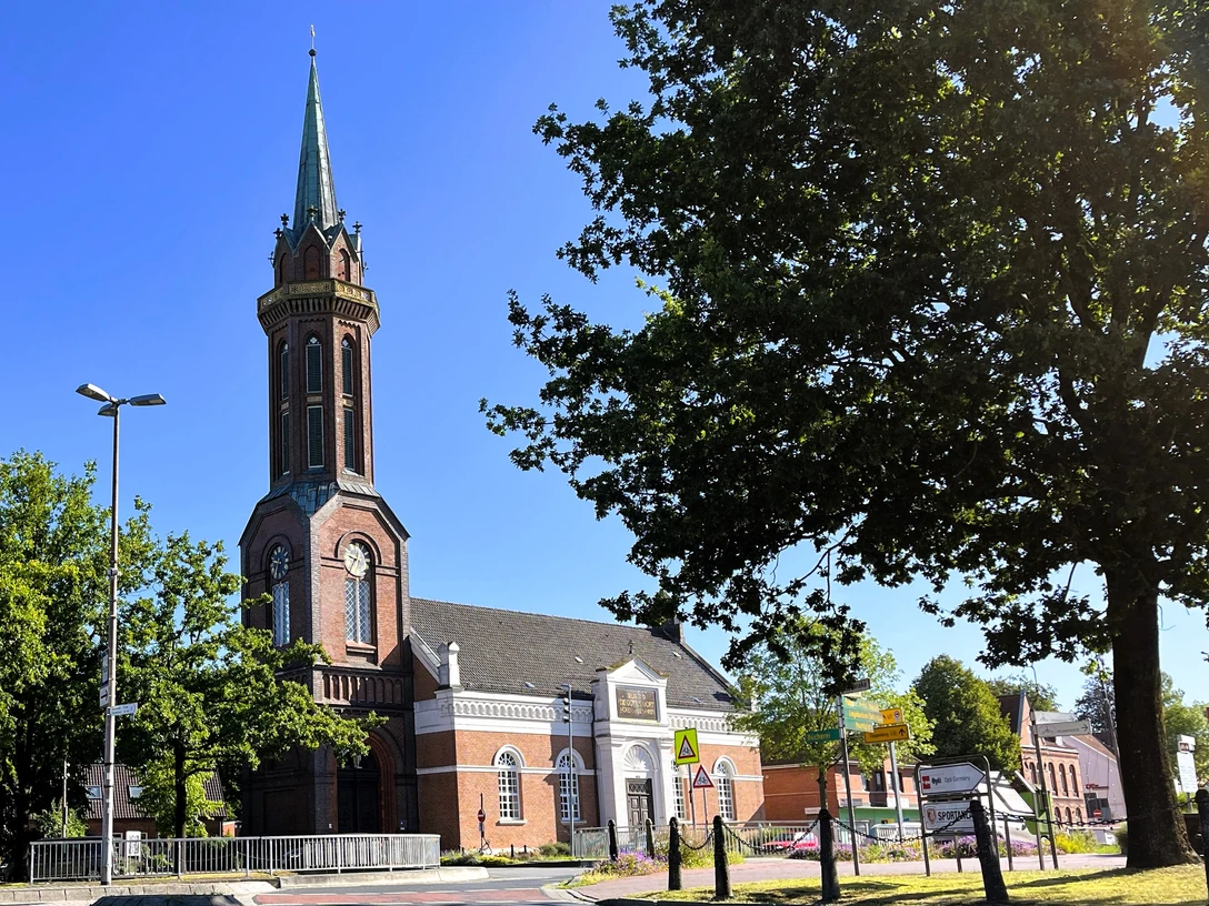 Hoffnungskirche Westrhauderfehn.JPG Kirchturm mit spitzem Dach neben Bäumen und einem Gebäude mit weißer Fassade unter blauem Himmel