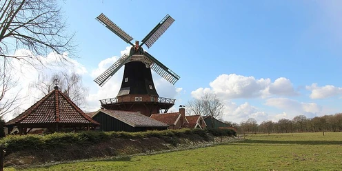 A windmill stands in a meadow under a cloudy sky with the sun low in the sky; on the left are trees with bare branches.