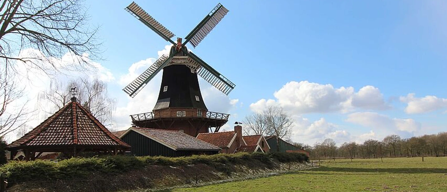 csm_Muehle_Rhaude-Rhauderfehn-Ostfriesland_13__93209e195e.jpg Windmühle steht auf einer Wiese unter bewölktem Himmel mit tiefstehender Sonne, links Bäume mit kahlen Ästen.