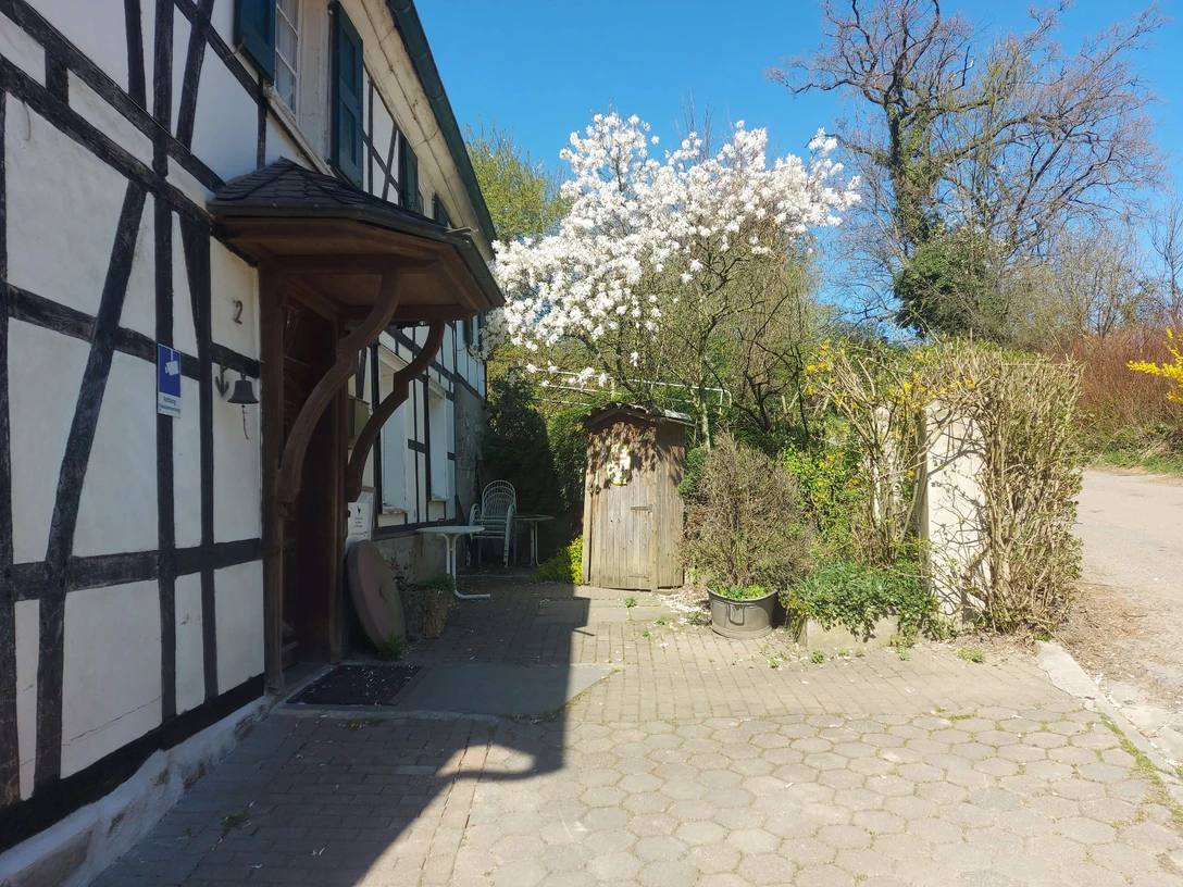 Gut Ehlenbeck in Haan Fachwerkhaus mit blühendem Baum vor der Tür, gepflasterter Weg und Gartenstühle unter blauem Himmel.