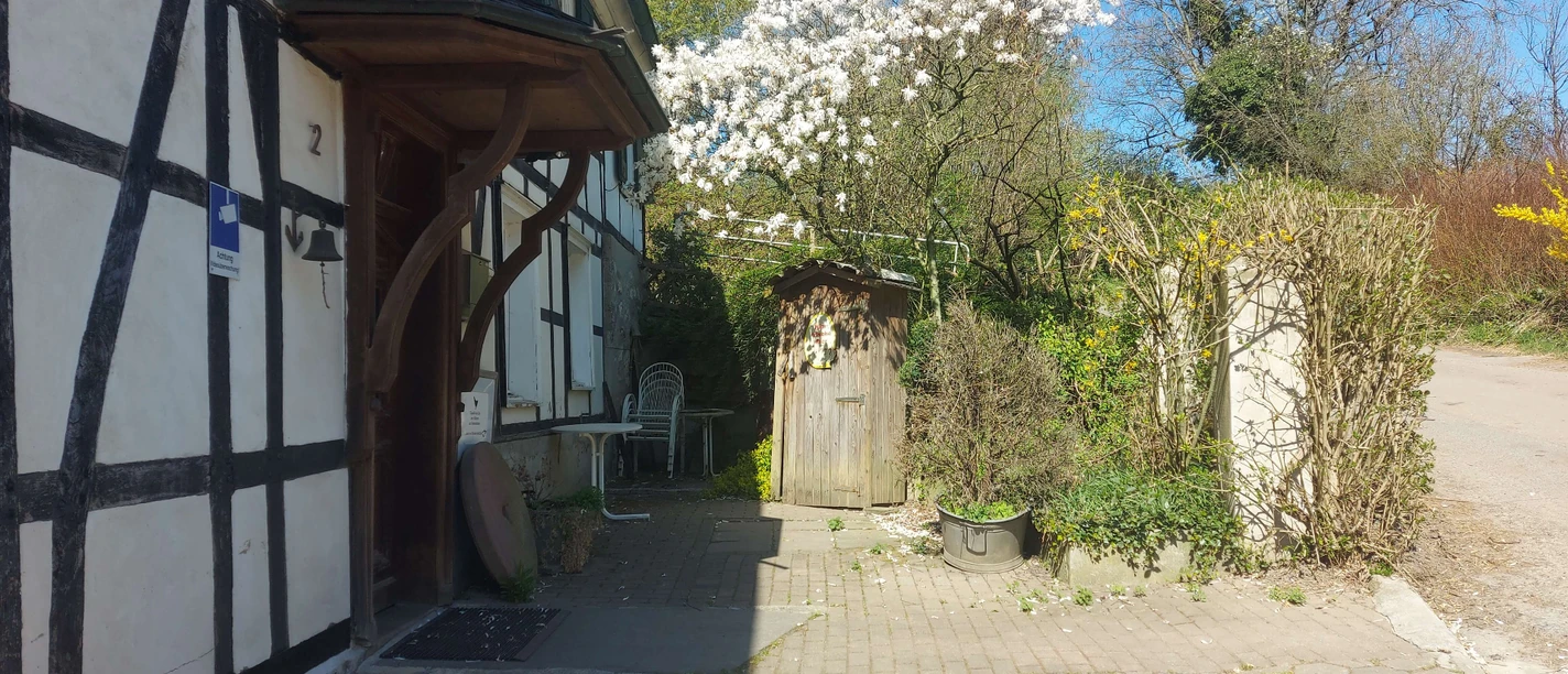 Gut Ehlenbeck in Haan Fachwerkhaus mit blühendem Baum vor der Tür, gepflasterter Weg und Gartenstühle unter blauem Himmel.