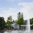 Eine große Springbrunnenfontäne in einem Park mit Blumenbeeten, Bäumen und einem weißen Pavillon.
