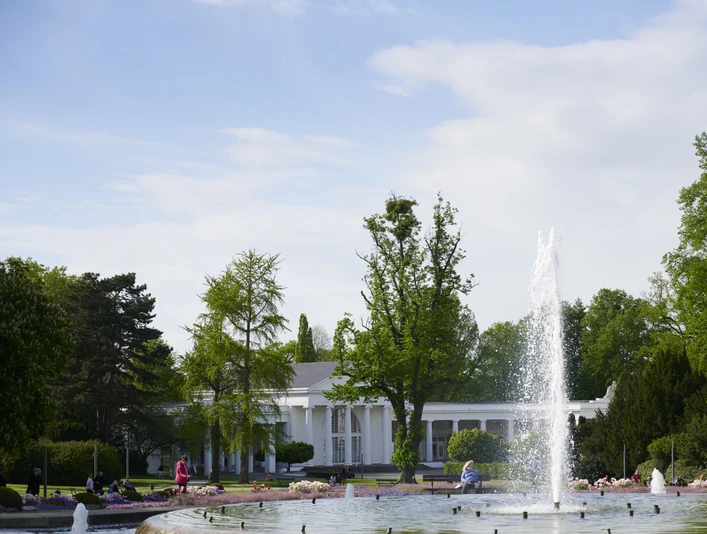 Mittelfontäne Eine große Springbrunnenfontäne in einem Park mit Blumenbeeten, Bäumen und einem weißen Pavillon.