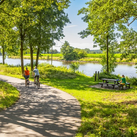 Zwei Radfahrende auf schattigem Weg entlang der Ems mit Picknickplatz unter grünen Bäumen.