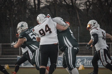 Spieler in Football-Ausrüstung im Zweikampf auf Spielfeld, dahinter Bäume.