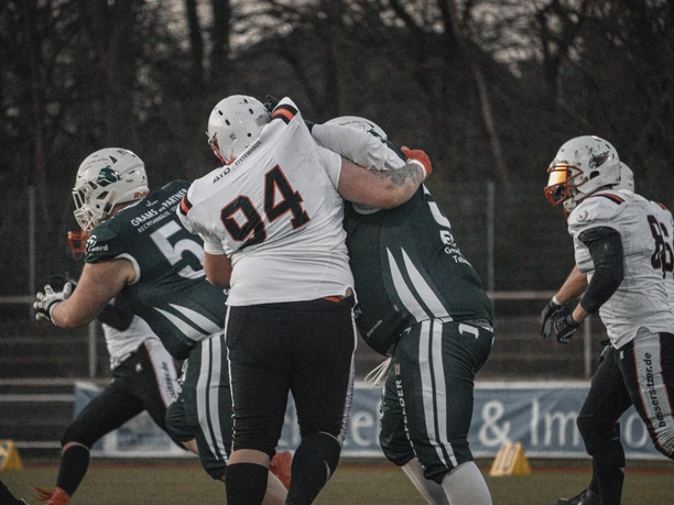 AFC Bielefeld Bulldogs Spieler in Football-Ausrüstung im Zweikampf auf Spielfeld, dahinter Bäume.