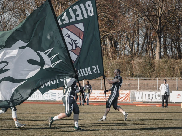 Flagge AFC Bielefeld Bulldogs American Football-Spieler halten große grüne Fahnen auf einem Spielfeld, Bäume im Hintergrund.