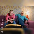 Zwei Männer sitzen in einem Retro-Wohnzimmer auf einem Sofa und schauen fern.