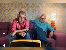 Zwei Männer sitzen in einem Retro-Wohnzimmer auf einem Sofa und schauen fern.