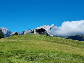 Holzhütte auf weiter Bergwiese vor hellen Felsgipfeln und einer vorbeiziehenden Wolke