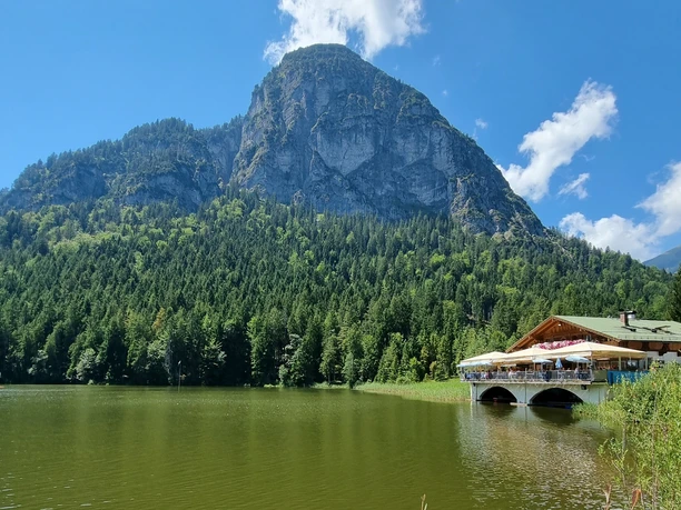 Berggasthof Pflegersee Berggasthof mit Seeterrasse vor bewaldetem Hang und markantem Felsmassiv unter klarem Himmel