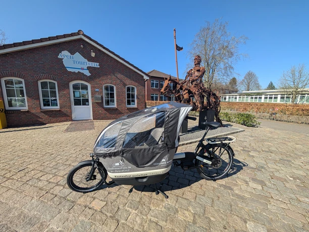 Lastenrad Ein Lastenrad, platziert auf einem gepflasterten Hof vor der Apen Touristik, daneben eine Skulptur einer historischen Figur.