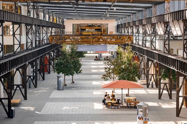 764_HS_OS_Campus_Lingen_Sep_2023-2.jpeg Innenraum der ehemaligen Werkshalle mit Stahlträgern, Bäumen, Sitzbereichen und Studierenden am Tisch