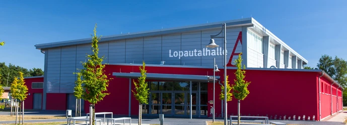 Außenansicht der modernen Lopautalhalle mit roter Fassade und grauem Dachbereich bei Sonnenschein.