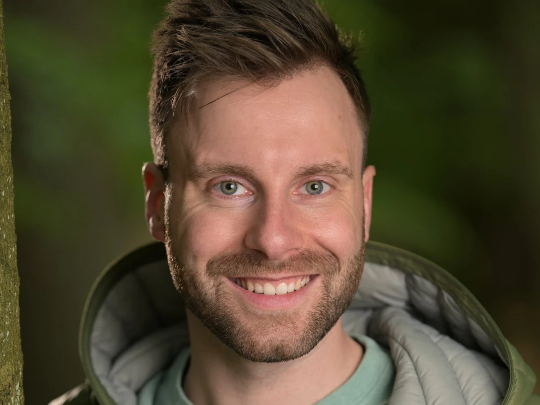 DSC_0911.JPG Lächelnder Mann in grüner Jacke vor unscharfem Waldhintergrund.