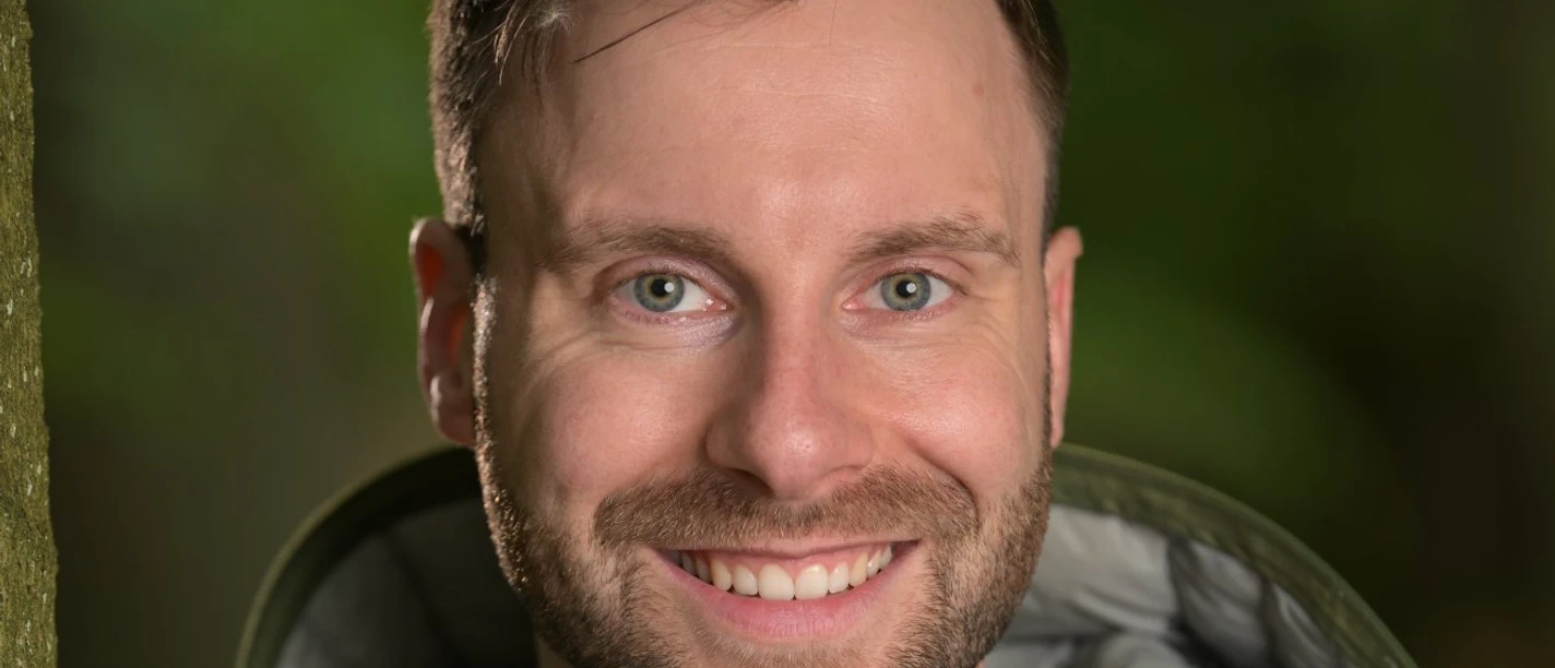 DSC_0911.JPG Lächelnder Mann in grüner Jacke vor unscharfem Waldhintergrund.