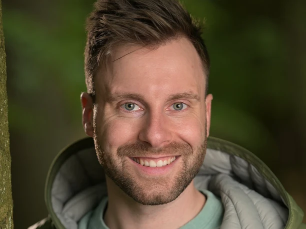 DSC_0911.JPG Lächelnder Mann in grüner Jacke vor unscharfem Waldhintergrund.