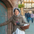 Historische Braumagd während einer Führung durch Einbeck Historische Braumagd mit Hand an der Klinke zum Rathauskeller und Körbchen im Arm während einer Führung durch Einbeck. Sie lächelt und im Hintergrund zeigen sich Passanten und ein Fachwerkgebäude.
