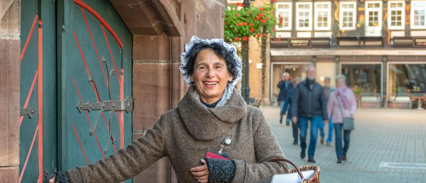 Historische Braumagd während einer Führung durch Einbeck Historische Braumagd mit Hand an der Klinke zum Rathauskeller und Körbchen im Arm während einer Führung durch Einbeck. Sie lächelt und im Hintergrund zeigen sich Passanten und ein Fachwerkgebäude.