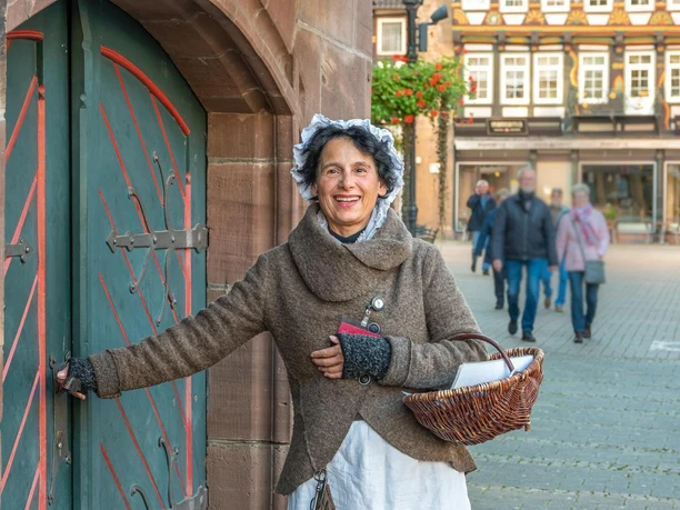 Historische Braumagd mit Hand an der Klinke zum Rathauskeller und Körbchen im Arm während einer Führung durch Einbeck. Sie lächelt und im Hintergrund zeigen sich Passanten und ein Fachwerkgebäude.
