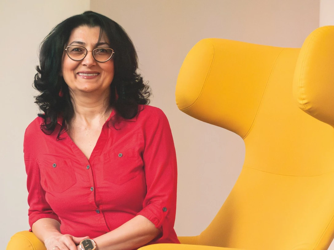 Ruzanna Shahinyan Ruzanna Shahinyan sitzt lächelnd neben einem großen, modernen gelben Sessel. Sie trägt eine rote Bluse.