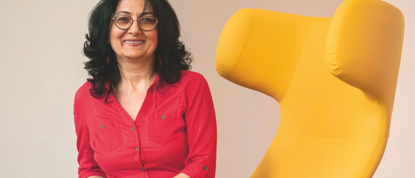 Ruzanna Shahinyan Ruzanna Shahinyan sitzt lächelnd neben einem großen, modernen gelben Sessel. Sie trägt eine rote Bluse.