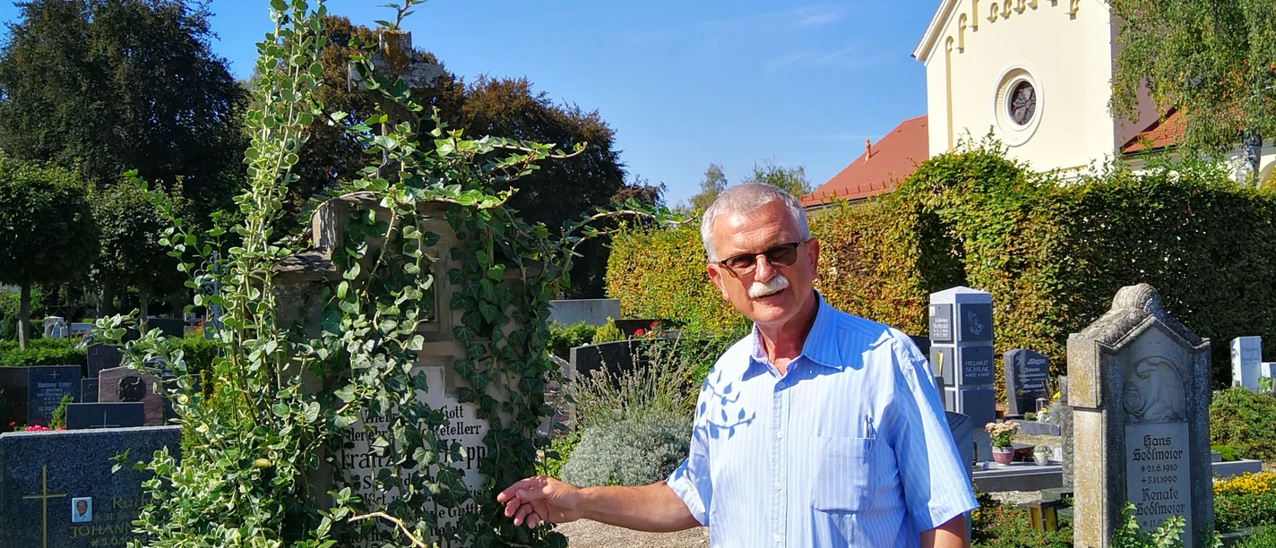 Ein Friedhof erzählt Auf dem Bild steht ein älterer Mann vor einem überwucherten Grabstein auf einem Friedhof.