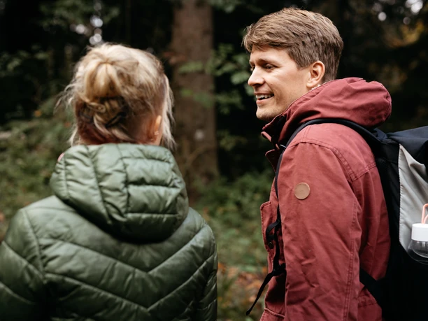 Een man en een vrouw wandelen in een bosrijke omgeving en dragen weerbestendige kleding.