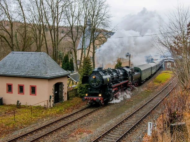 A historic steam train passes an idyllic station surrounded by nature in the fall