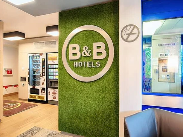 Eine moderne Hotel-Lobby mit einem grünen B&B Hotels-Schild. Der Empfangsbereich ist hell und einladend gestaltet.