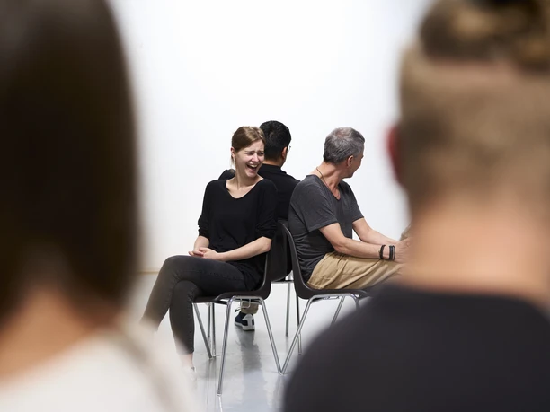 Workshop Team IMPROve Menschen auf Stühlen, Rücken an Rücken, im Dialog. Frau lächelnd, entspannte Gesprächsatmosphäre.