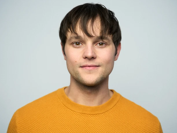 Coach Carlo Woltiri Ein junger Mann mit braunen Haaren und einem gelben Pullover auf neutralem Hintergrund.