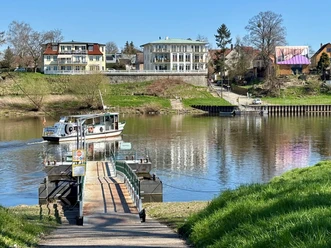 Fähre F10 zwischen Birkwitz und Heidenau Ein kleines Fährboot überquert einen ruhigen Fluss, im Hintergrund stehen bunte Gebäude und Bäume unter einem klaren blauen Himmel.