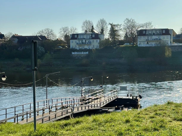 Fähreanleger F10 Heidenau Ein Holzsteg führt über einen Fluss, im Hintergrund sind zwei mehrstöckige Gebäude und Bäume zu sehen, bei klarem Himmel und ruhiger Atmosphäre.