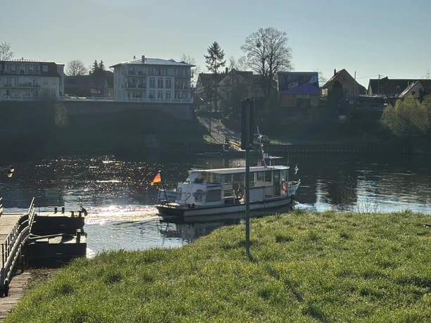 Een klein passagiersschip onder Duitse vlag vaart op een rivier, omringd door een groene weide en huizen op de achtergrond onder een heldere hemel.